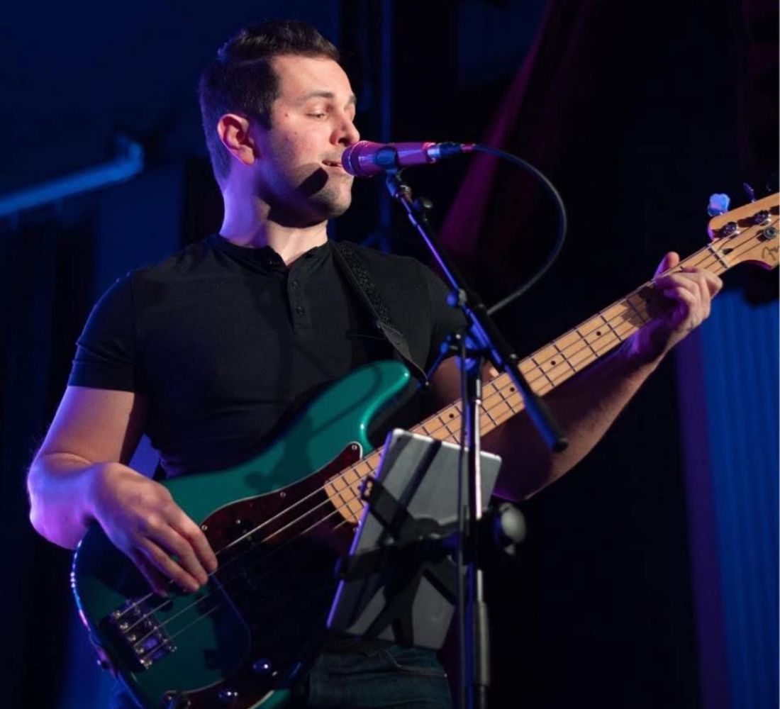 A Straight A’s band member playing a teal bass guitar at the microphone on stage under blue lighting