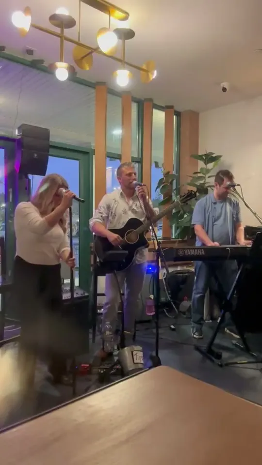 A Straight A's vocalist, acoustic guitarist, and keyboardist performing in front of tall windows at a venue